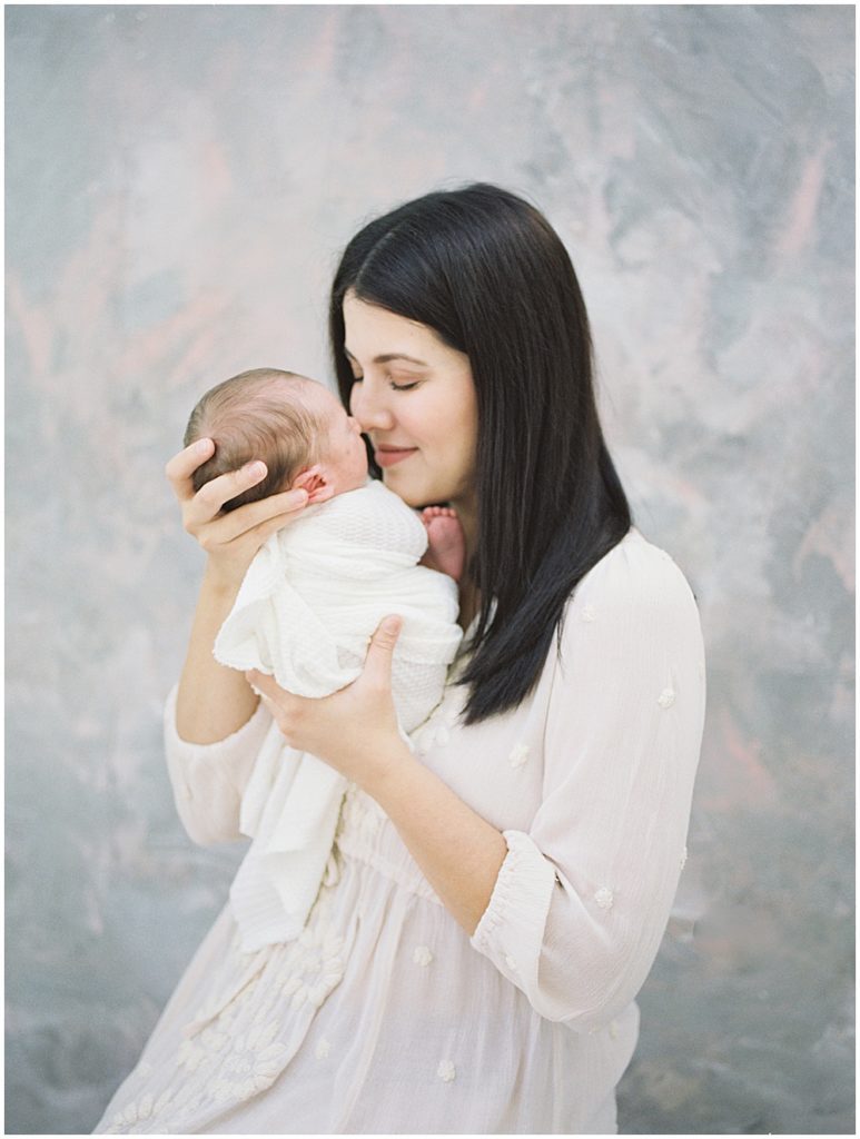 Mother With Brown Hair And Pink Dress Brings Newborn Baby Up To Her Nose And Closes Her Eyes During Studio Newborn Session.