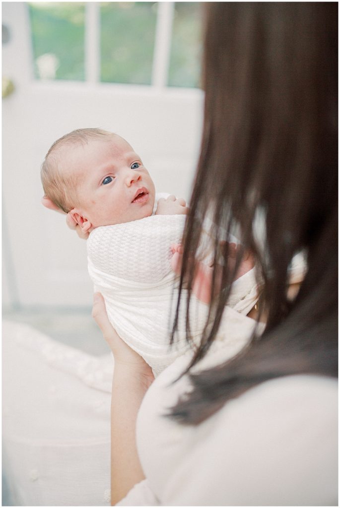 Newborn Baby Boy Is Held In Front Of His Mother And Looks Up At Her With Big Blue Eyes.