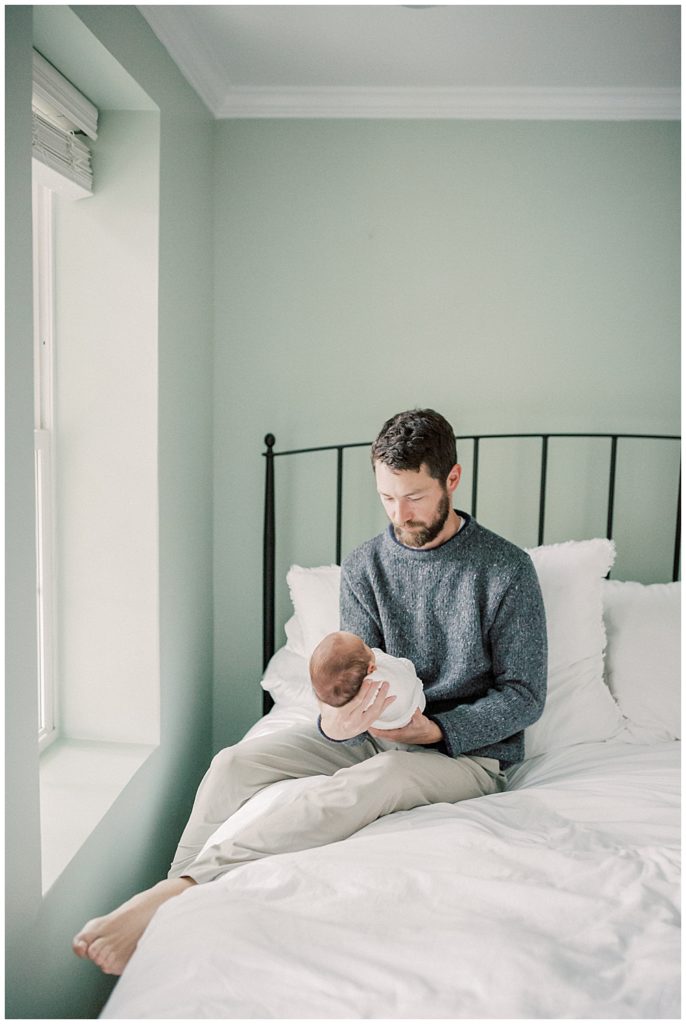 Father With Beard And Gray Sweater Sits On Bed Holding Newborn In Front Of Him During Dc Row House Newborn Session.