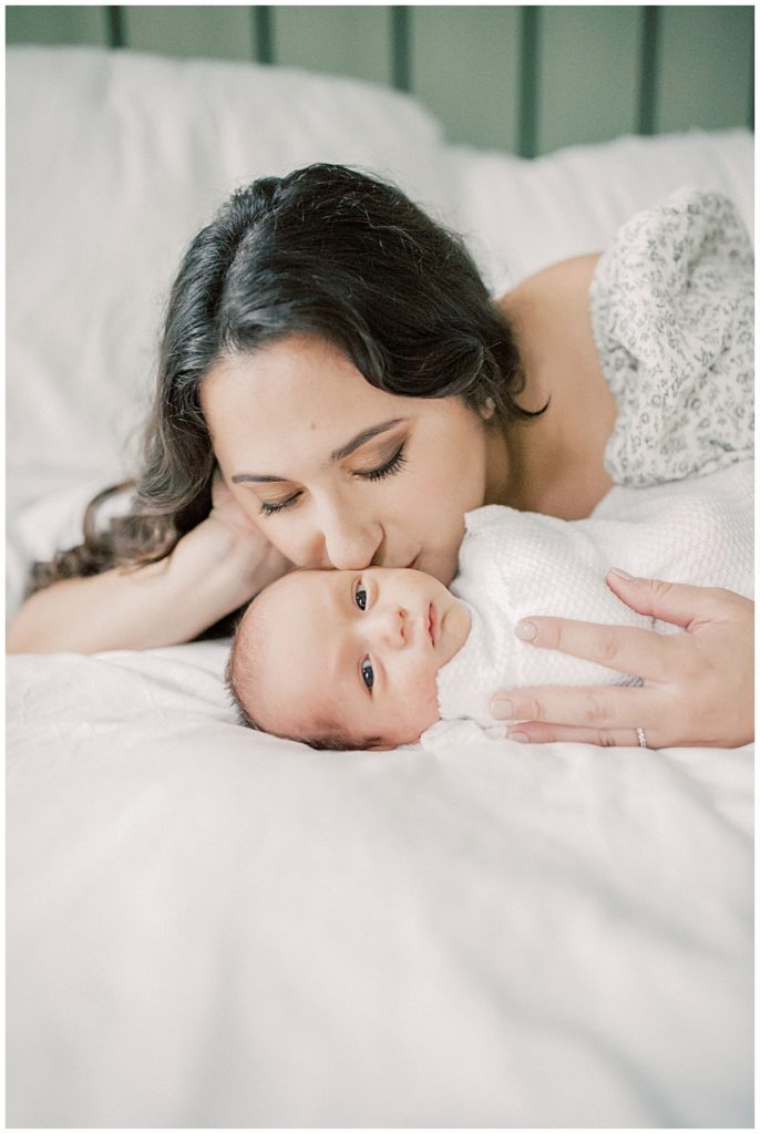 Mother Kisses Newborn Baby On Bed As Baby Looks At The Camera.