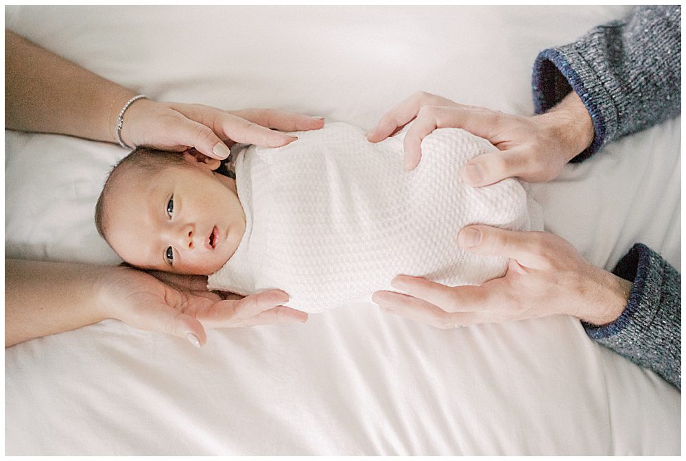 Baby Swaddle In White Is Held On Bed By Mother And Father.