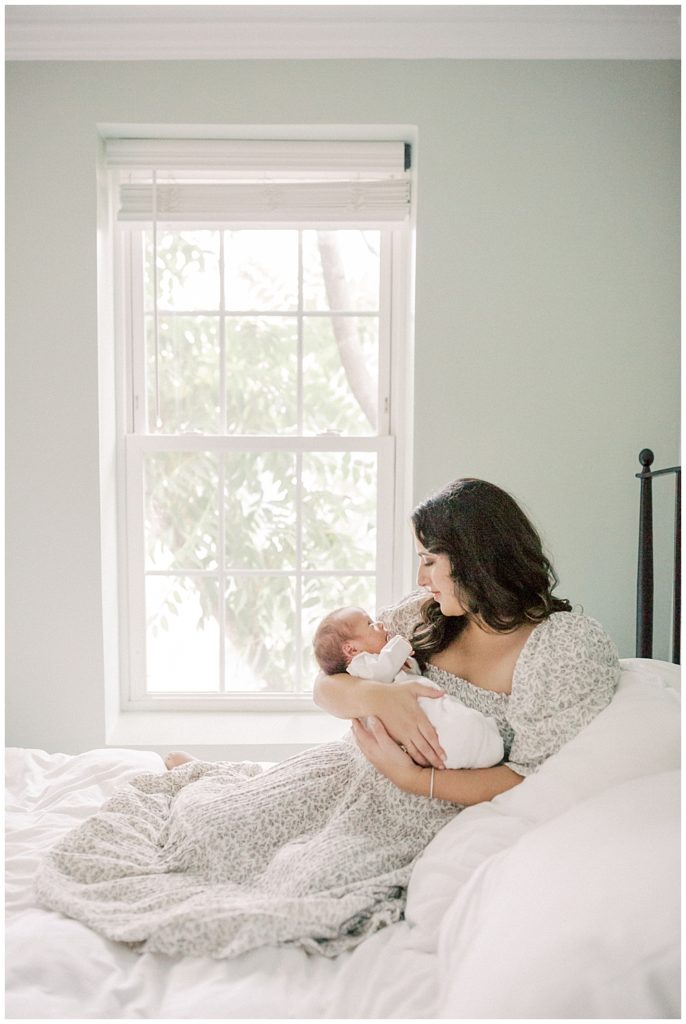 Mother Sits On Bed By Window Holding Newborn During Dc Row House Newborn Session.