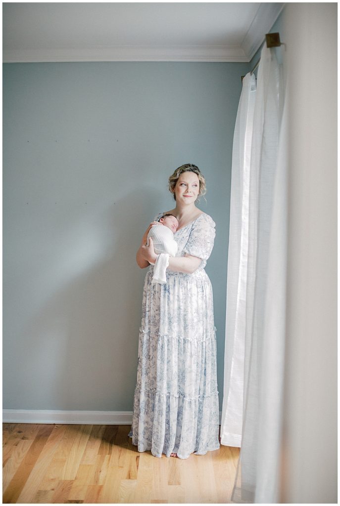 Mother Looks Out The Window With Soft Smile While Wearing A Blue Dress Holding Her Newborn During Fairfax Va Newborn Session.