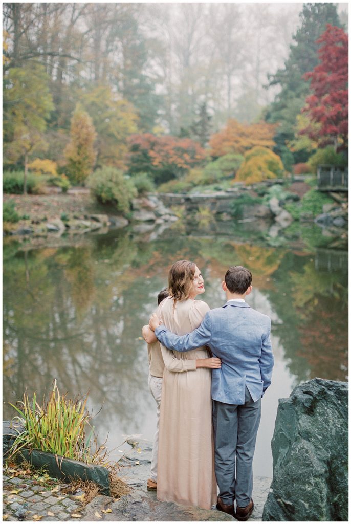 A Mother Embraces Her Two Boys At The End Of A Pond And Looks Over Her Shoulder At The Camera Family Session With Older Children.