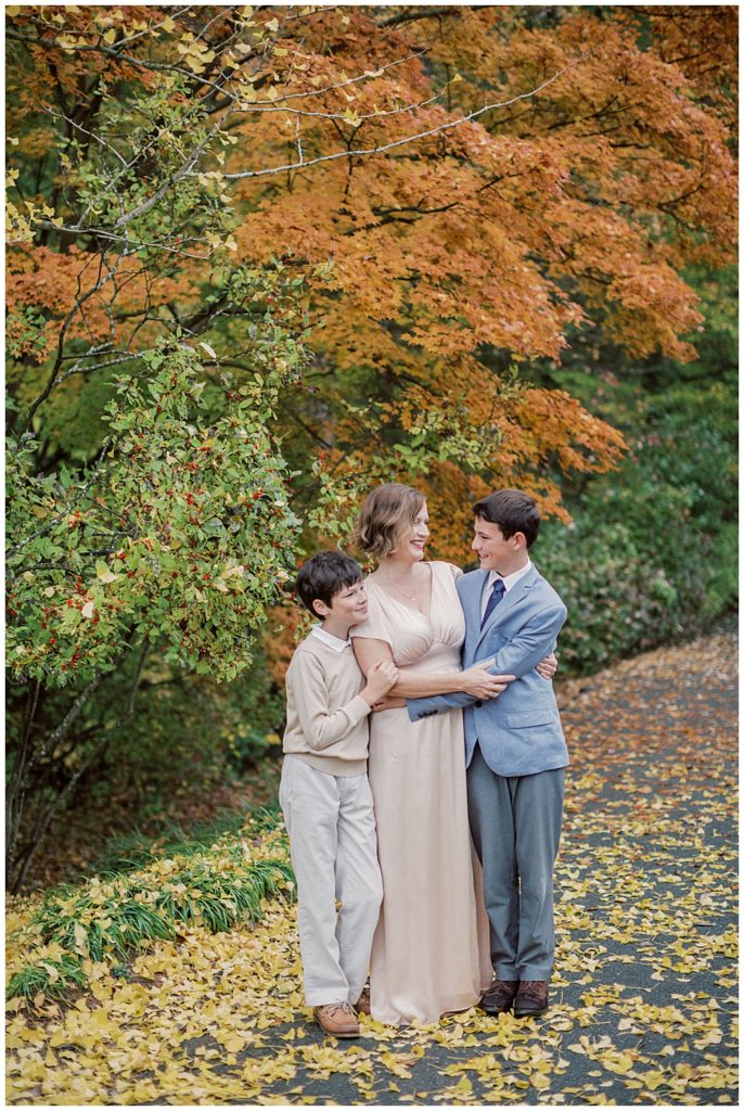 Mother Embraces Her Two Older Sons During Their Family Session With Older Children On A Fall Day At Brookside Gardens In Maryland.