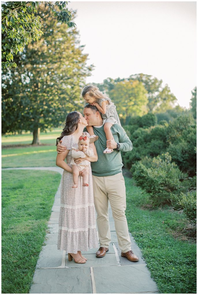 Mother And Father Hold Their Two Daughter While Leaning In For A Kiss During Glenview Mansion Photos By Marie Elizabeth Photography.