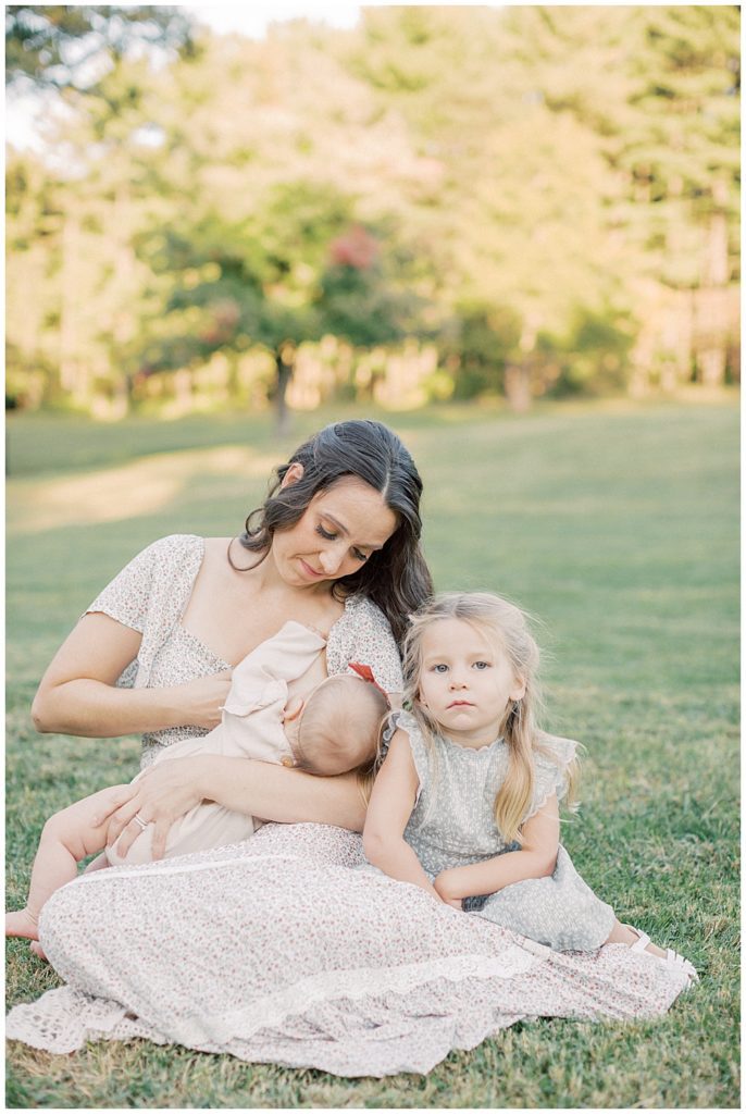 Mother Nurses Her Infant Daughter While Sitting In A Field With Her Toddler Daughter Sitting Next To Her.