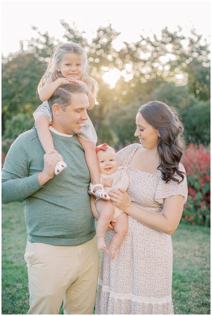 Mother And Father Hold Their Two Daughters All Smiling While Standing In Front Of Red Flowers During Glenview Mansion Photos By Marie Elizabeth Photography.