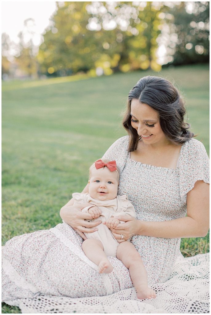 Baby Girl Smiles While Sitting On The Lap Of Her Mother In Floral Dress.