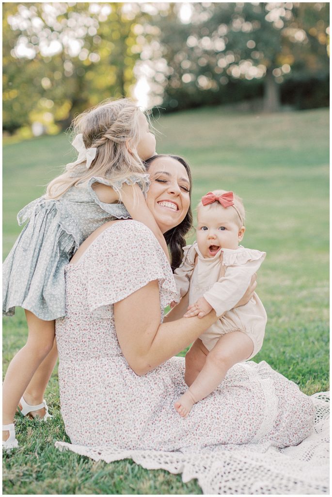 Toddler Girl Comes Over And Gives Mother A Hug While She Holds Her Baby Girl As They Sit And Laugh In A Field.