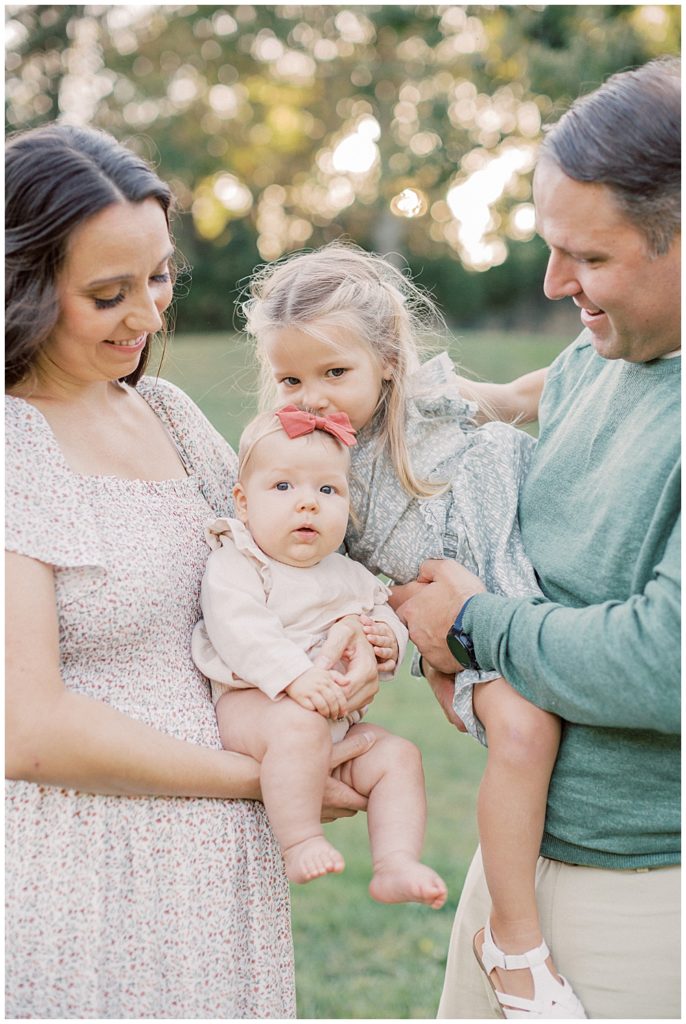 Father Holds Toddler Daughter As She Leans In To Kiss Her Baby Sister Held By Mother During Glenview Mansion Photos By Marie Elizabeth Photography.