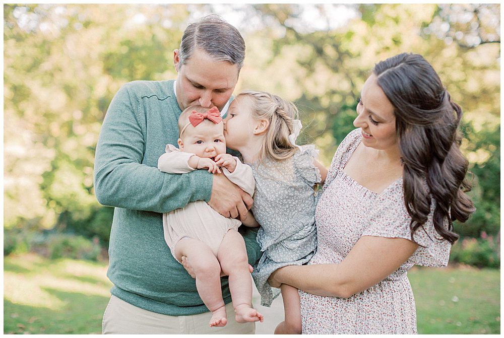 Big Sister Held By Her Mother Leans Over And Kisses Her Baby Sister On The Head During Glenview Mansion Photos By Marie Elizabeth Photography.