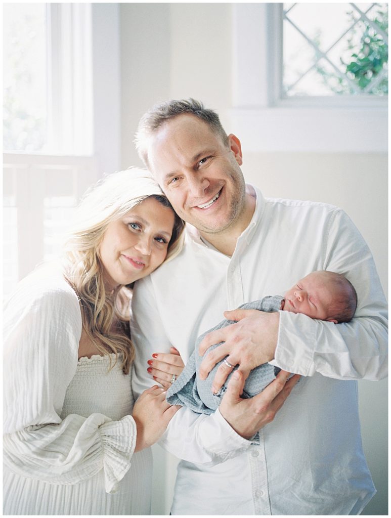 Mother Rests Her Head On Husband's Shoulder As He Holds Their Newborn Son.