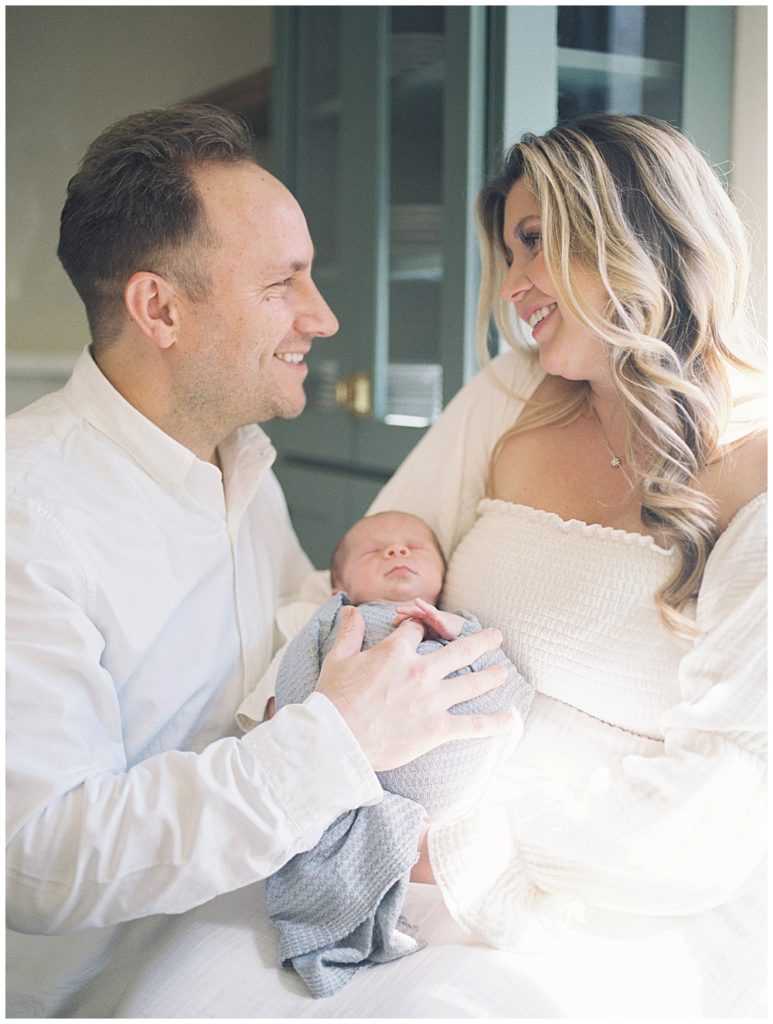 Father And Mother Look At One Another And Smile While Holding Their Baby Boy In Their Kitchen In Washington, Dc.