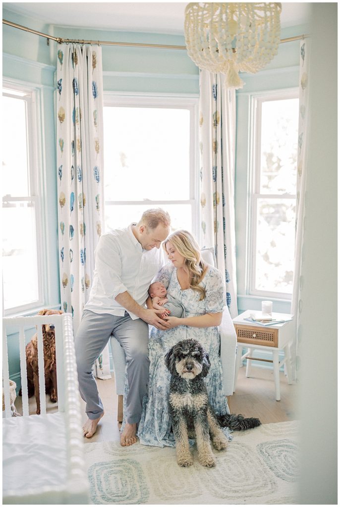 Father And Mother Sit On Chair In Nursery Holding Son During Newborn Session With Dogs In Washington, Dc.