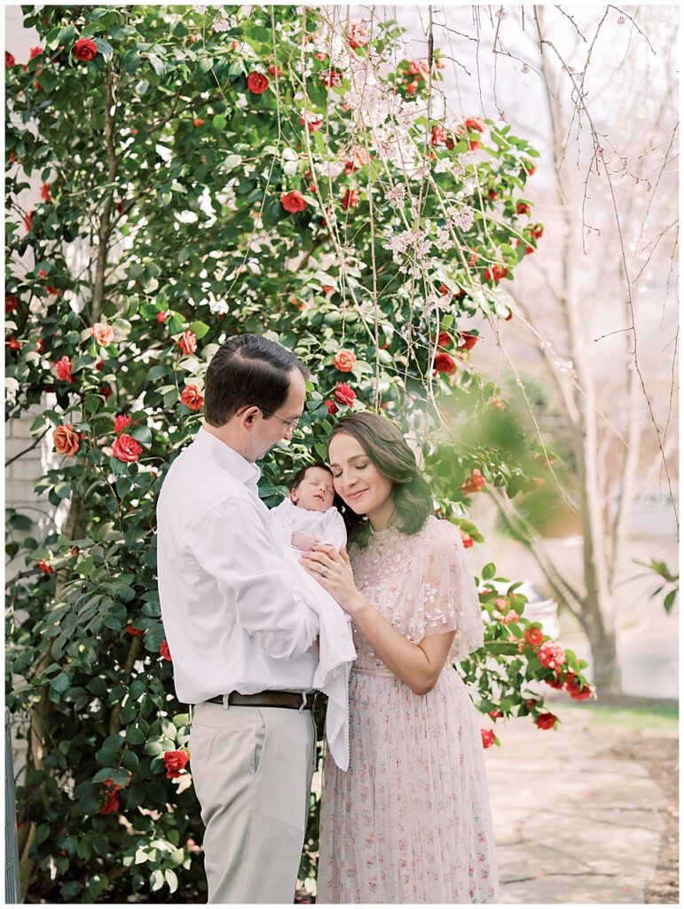 Mother In Pink Needle &Amp; Thread Dress Leans Into Her Baby Girl Held By Her Husband As They Stand In Front Of A Camellia Bush Photographed By Arlington Newborn Photographer Marie Elizabeth Photography.