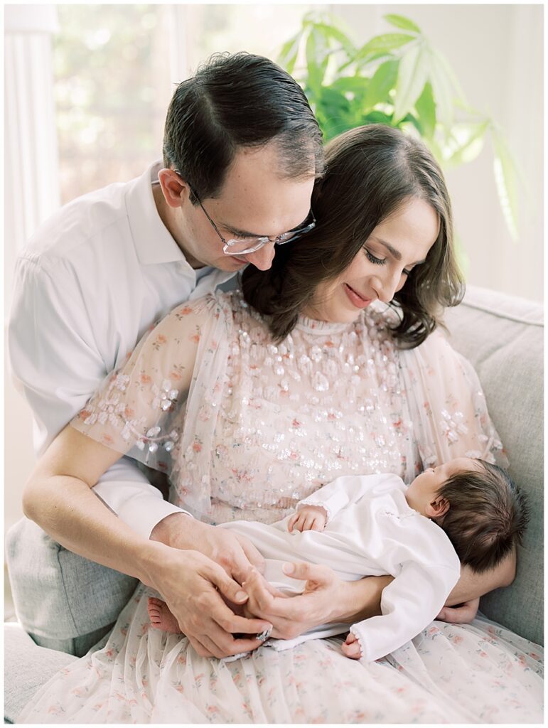 Mother And Father Cuddle Together And Look Down At Their Baby Girl While Sitting On Their Couch.