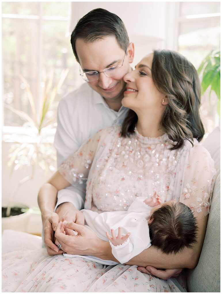 Mother And Father Lean Into One Another In A Cuddle As They Hold Their Baby Girl On Their Couch.