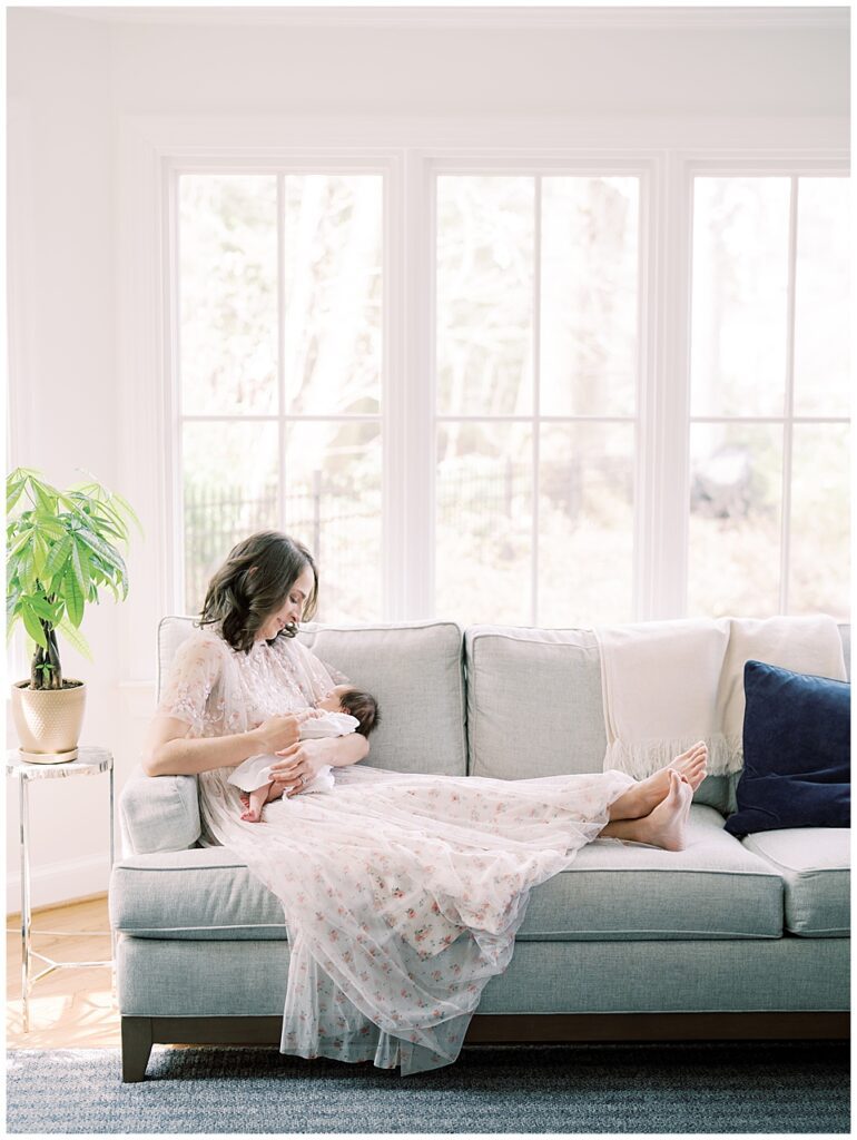 Brown-Haired Mother Sits On Her Couch, Gazing Down At Her Baby Photographed By Arlington Newborn Photographer Marie Elizabeth Photography.