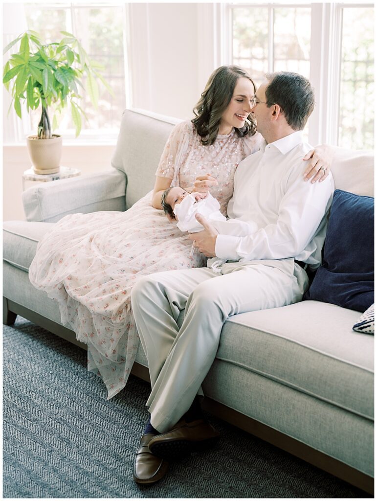 Mother And Father Sit On The Couch With Baby And Lean Into One Another.