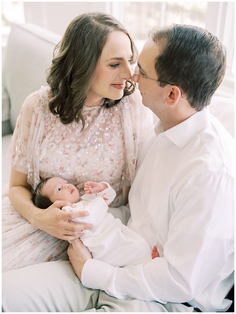 Mother And Father Lean Into One Another As They Hold Their Baby Girl On Their Couch During Their Arlington Newborn Session.