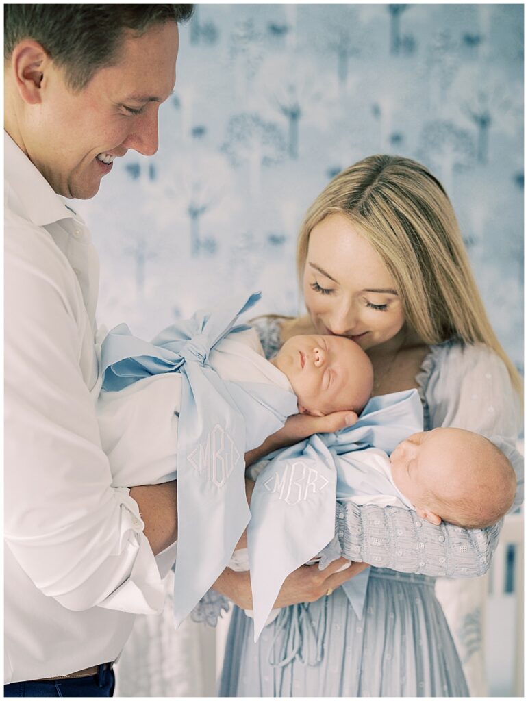 Mother Leans Down To Kiss One Of Her Twin Babies Wearing A Beaufort Bonnet Bow Swaddle As Her Husband Holds Him While She Holds The Other Baby In Their Serena &Amp; Lily Nursery.