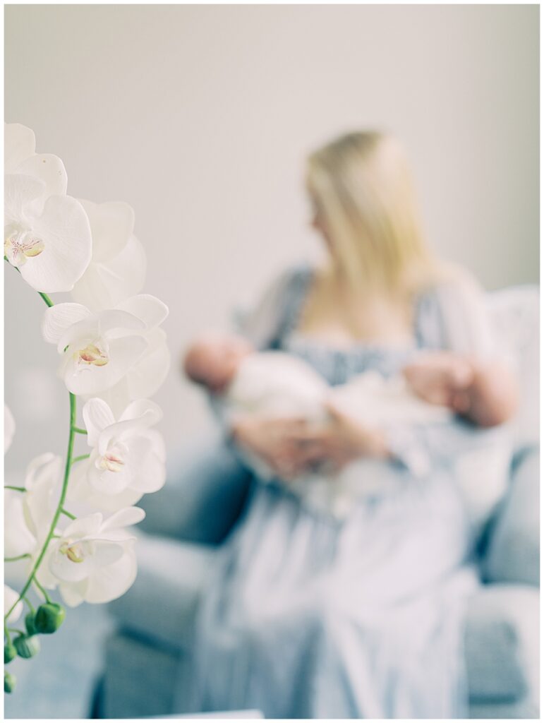 A White Orchid Can Be Seen In The Foreground While A Blond Mother Wearing A Blue Dress Holding Twin Baby Boys Sits In The Background, Photographed By Newborn Photographer Marie Elizabeth Photography.