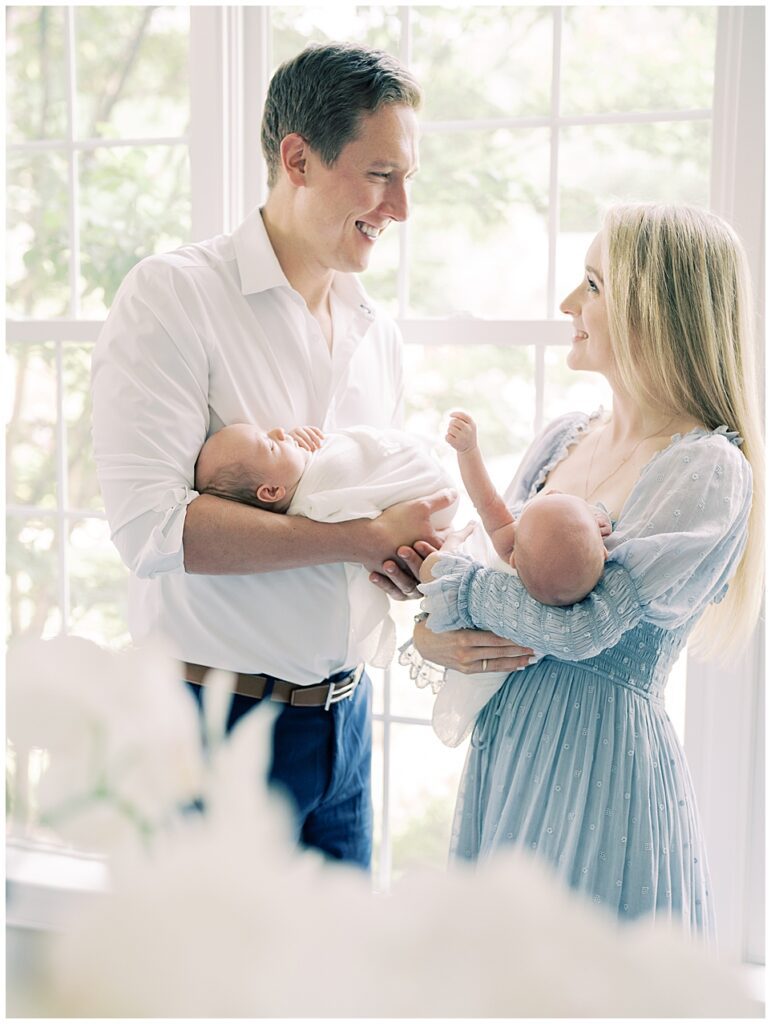 Blonde Mother And Father Smile At One Another While Standing In Front Of A Window During Their Chevy Chase Newborn Session.