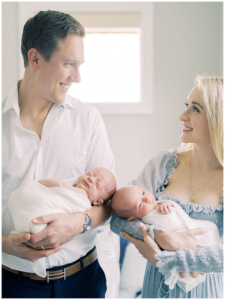 A Mother And Father Hold Their Twin Boys Swaddled In White During Their Chevy Chase Newborn Session.