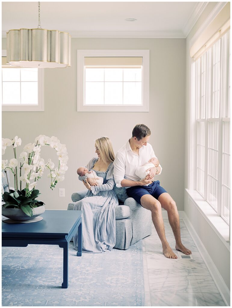 Father And Mother Sit On A Chair In Their Living Room Looking Down At Their Twin Baby Boys, Photographed By Newborn Photographer Marie Elizabeth Photography.