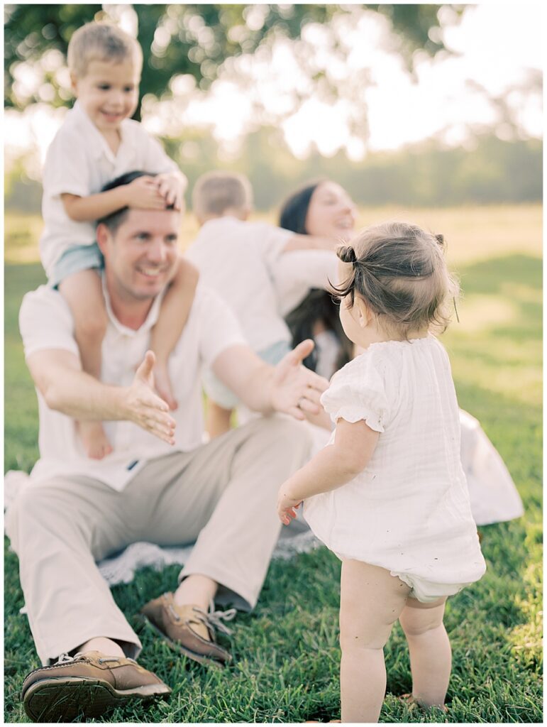 Father Stretches Hands Out To Toddler Daughter As He Sits With His Sons And Wife During Manassas Battlefield Family Session.