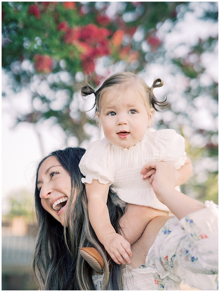 Little Girl In Pig Tails Sits On Her Mother's Shoulders As Her Mother Smiles.