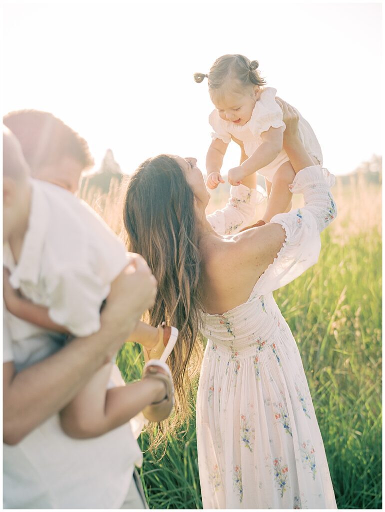 Mother Holds Up Toddler Daughter In The Air As Father Holds Toddler Son In The Foreground.