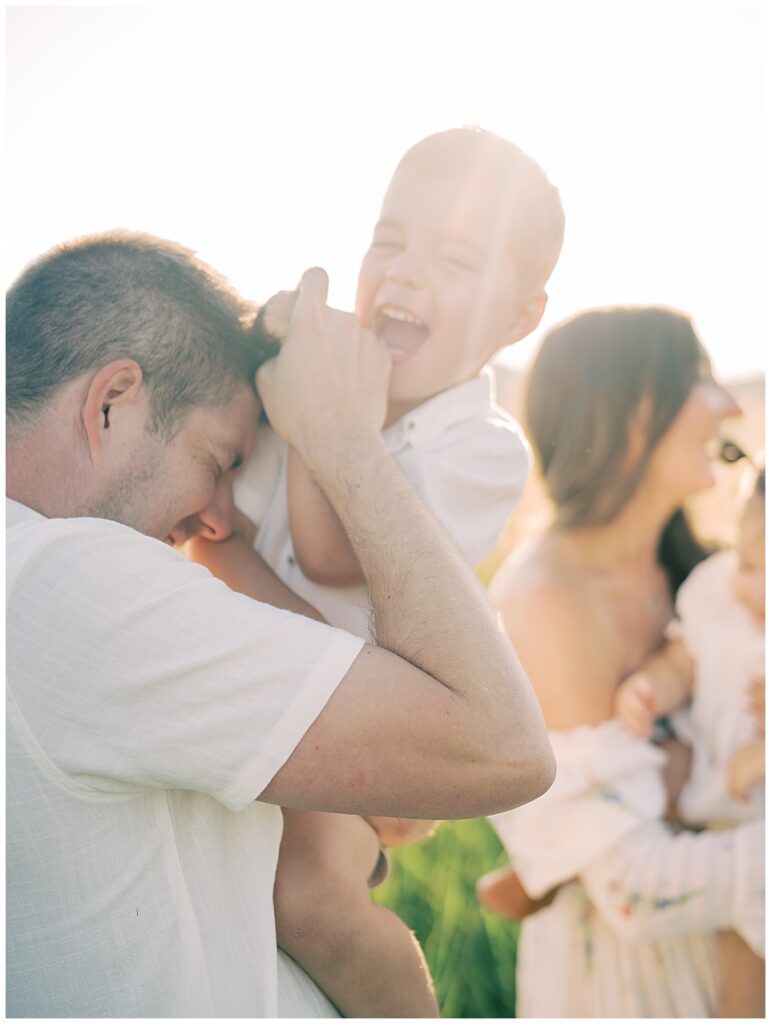 Little Boy Held Up By His Father Laughing During Sunrise Family Session At Manassas Battlefield.