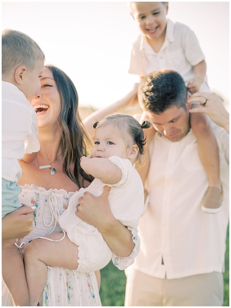 Mother With Brown Hair Smiles At Her Son As She Holds Her Toddler Daughter And Stands With Her Husband Who Has Toddler Son On His Shoulders.