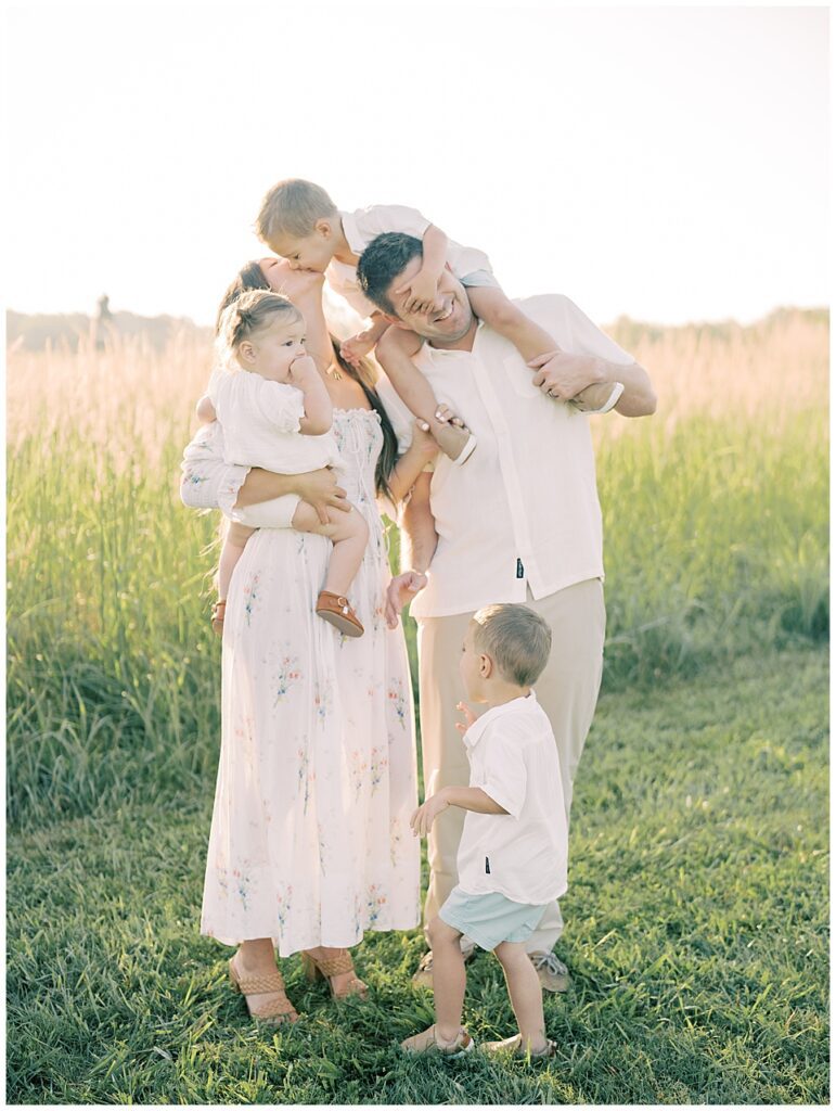 Little Boy On Father's Shoulders Leans Down To Kiss His Mother During Manassas Battlefield Family Session At Sunrise.