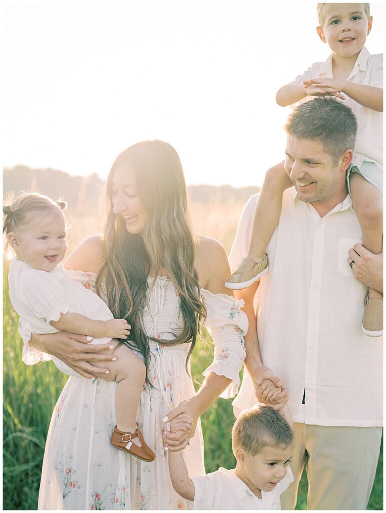 Mother And Father Smile At Daughter As They Stand With Three Children At Sunrise During Manassas Battlefield Family Session.