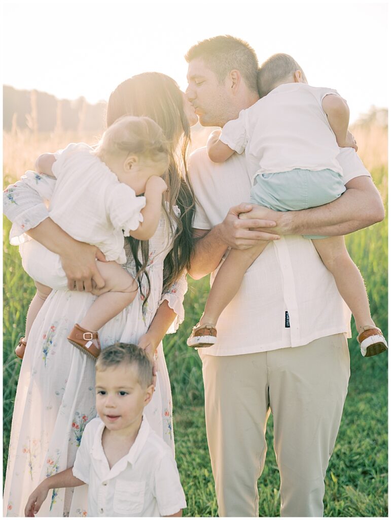 Mother And Father Kiss While Holding Their Two Children And Standing With Son During Manassas Battlefield Family Session.