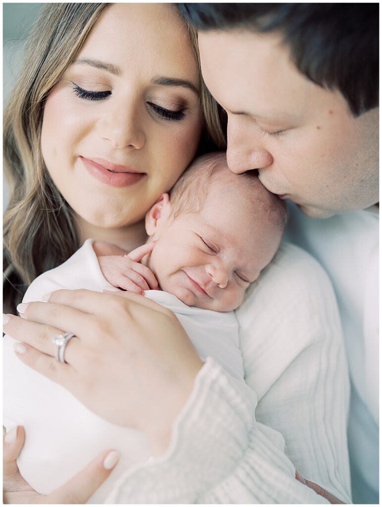 A Newborn Baby Swaddled In White Smiles As She Lays On Her Mother's Shoulder While Her Father Kisses Her Head During Their North Bethesda Newborn Session.