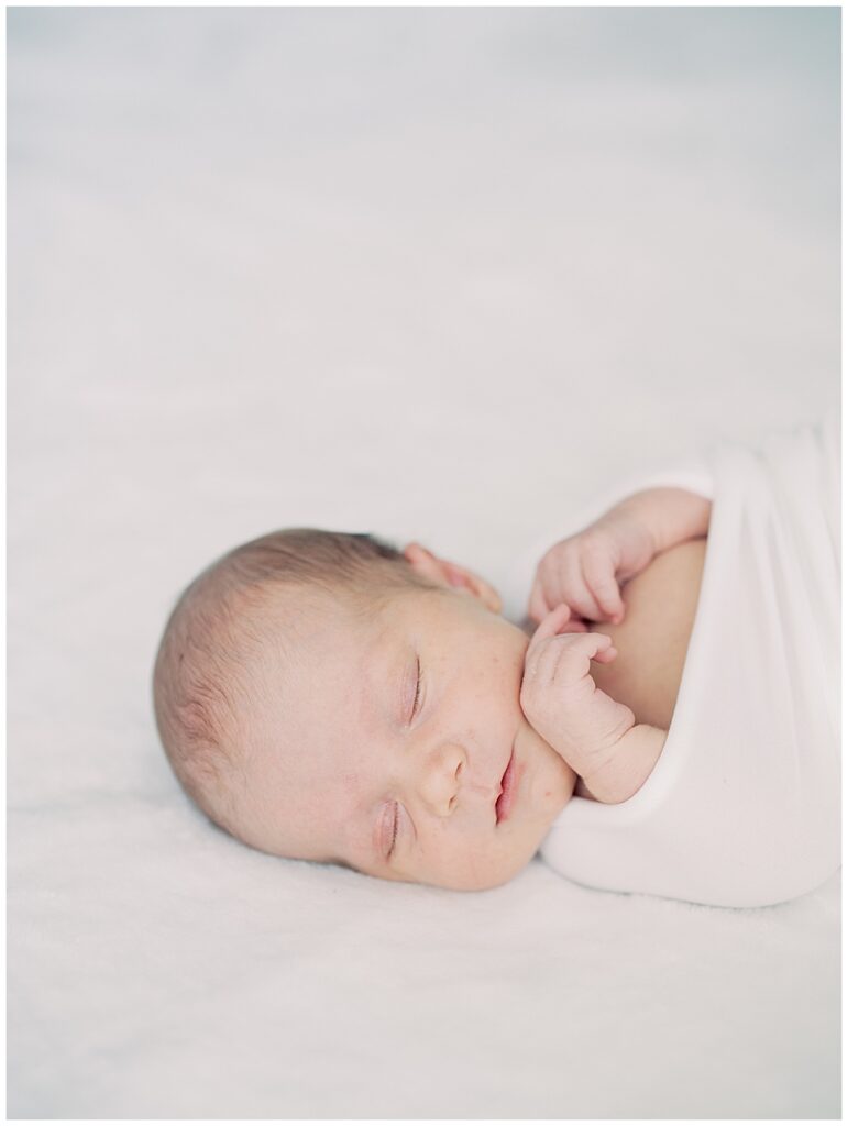 Baby Swaddled In White Sleeps With Hands Up By Her Face.