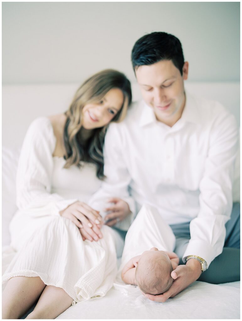 Image Of Mother Leaning Her Head On Her Husband's Shoulder While They Sit On The Bed Admiring Their Newborn Daughter Sleeping In Front Of Them.