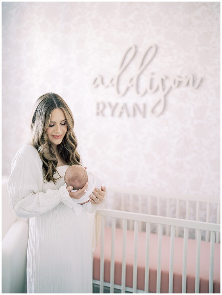 Mother With Long Brown Hair Holds Her Baby Girl In Front Of Her In Their Pink Nursery During Her North Bethesda Newborn Session.