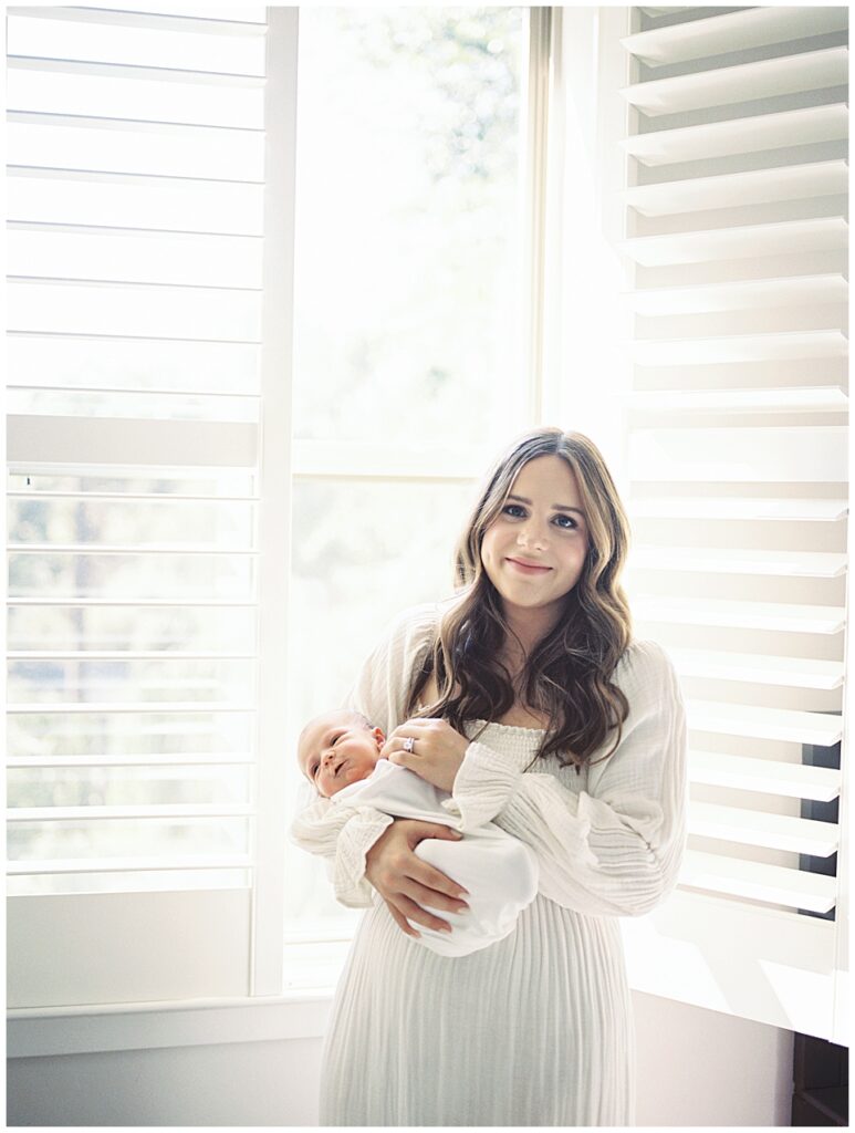 Long Brown-Haired Mother Holds Her Newborn Baby Girl While Standing In Front Of Blinds Holding Baby Girl During Her North Bethesda Newborn Session.