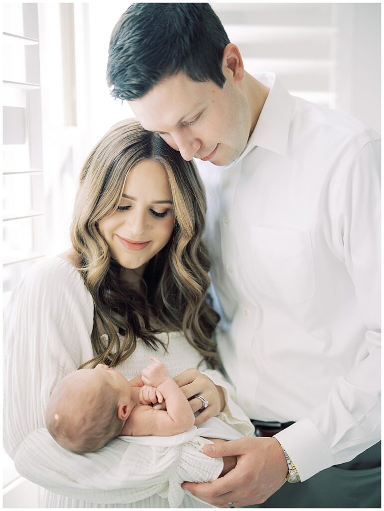 Mother And Father Stand Huddled Together Holding Their Baby Girl During Her North Bethesda Newborn Session.