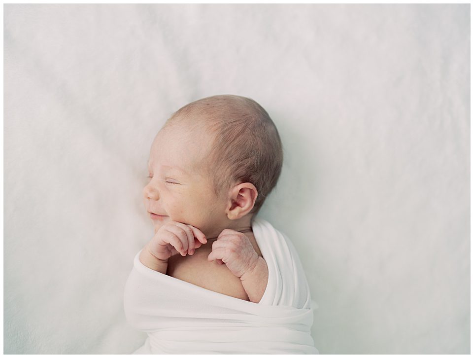 Newborn Baby Swaddled In White Smiles As She Lays On A White Backdrop During Her North Bethesda Newborn Session.