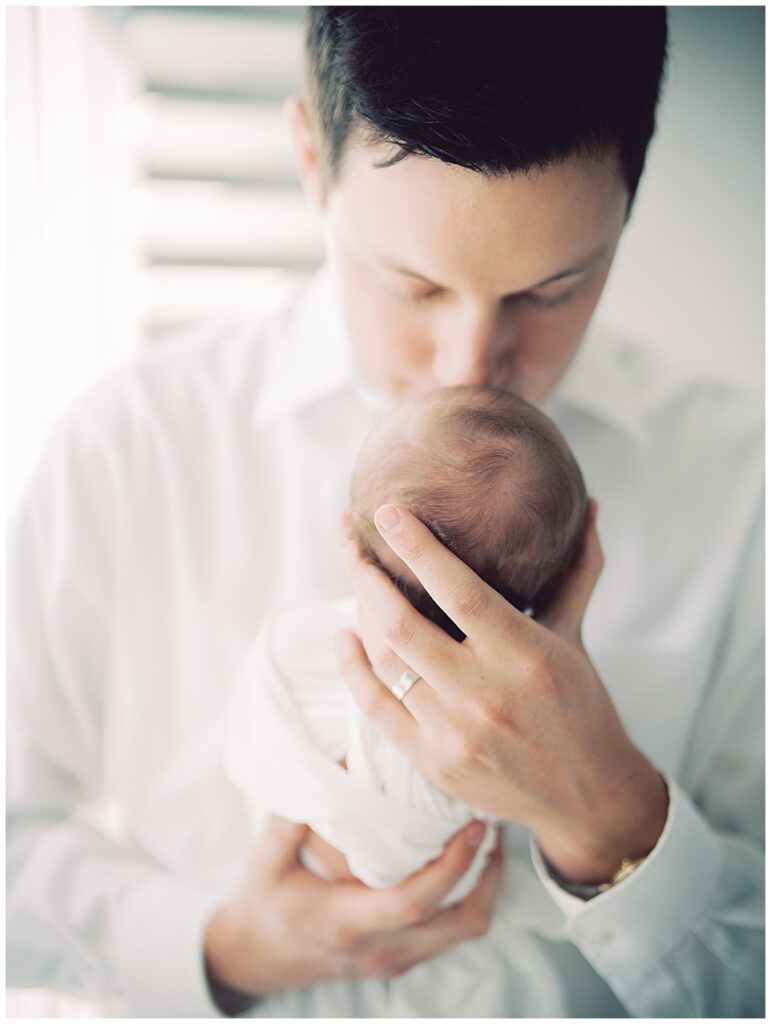 Brown Haired Father In White Shirt Brings Up Baby Girl To Kiss Her On The Head.