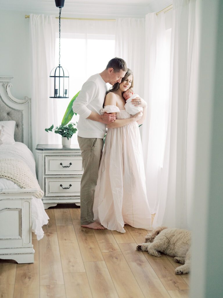 Mother And Father Stand Cuddled Together Holding Baby With Dog Laying Nearby During Their Newborn Photos In Annapolis.