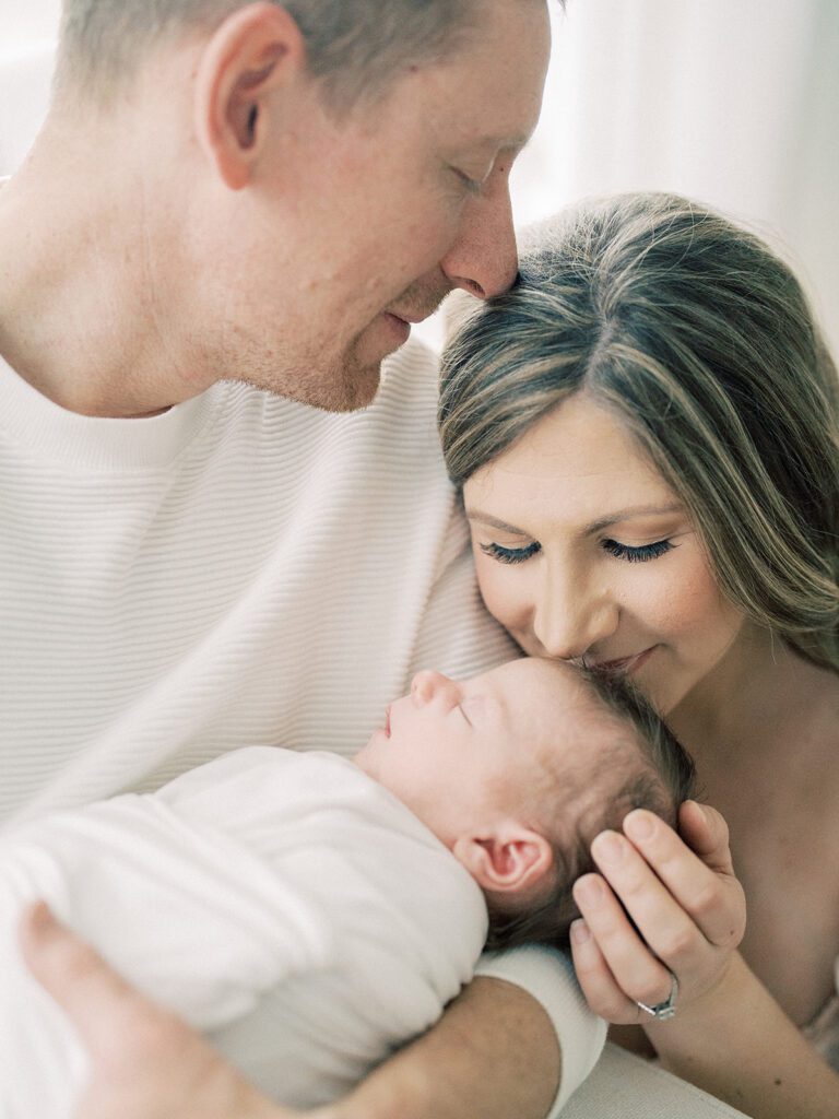 Mother Kisses The Top Of Baby's Head As Husband Leans Into Her As He Hold Their Newborn Baby Swaddled In White.