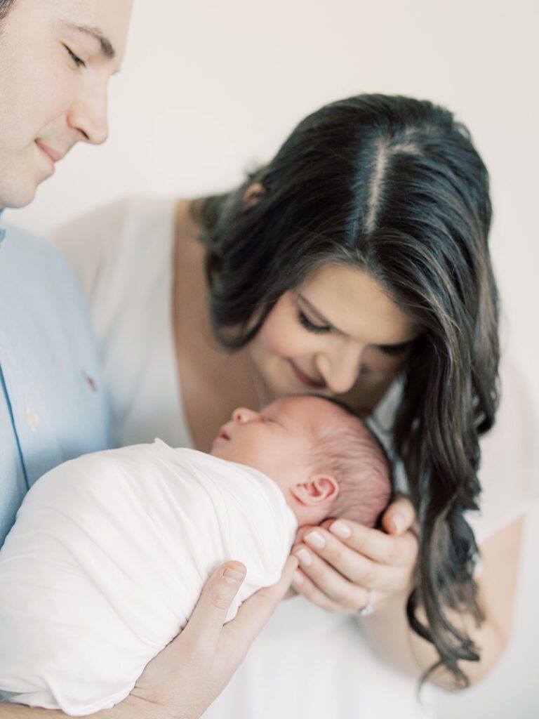 Mother With Long Brown Hair Leans Down To Kiss The Top Of Her Baby's Head As Her Husband Holds Baby During Their Georgetown Newborn Session.