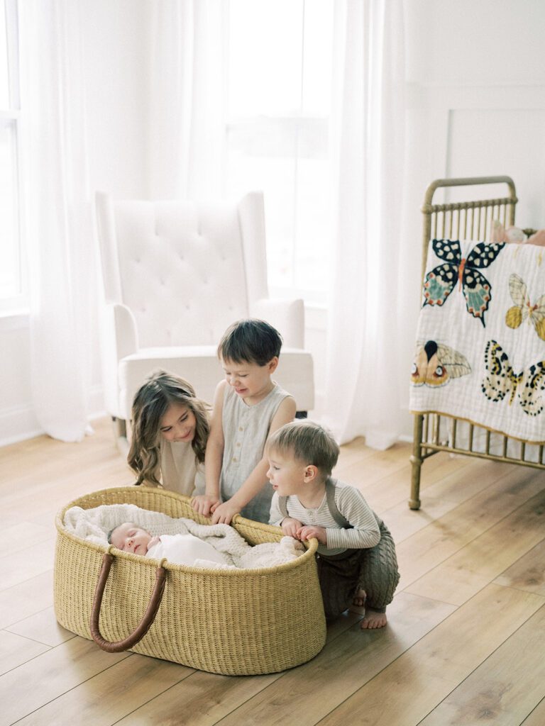 Three Toddler Siblings Lean Into A Moses Basket Sitting In A Nursery To Admire Their Newborn Baby Sister.